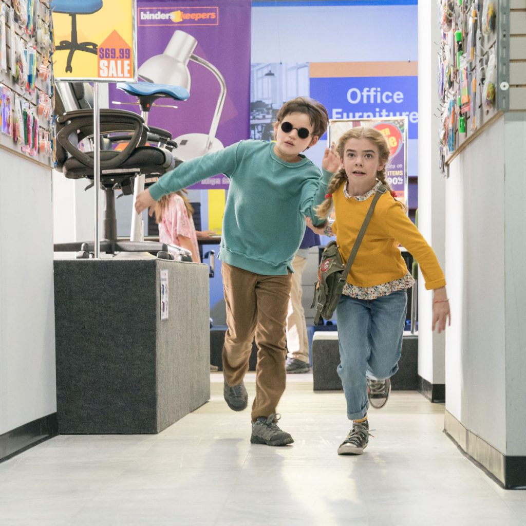 Dos niños corriendo en una tienda, el niño tiene lentes de sol y la niña una bolsa colgada de lado