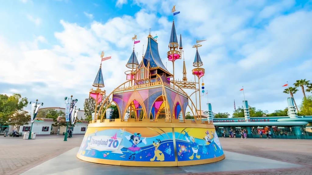 Una escultura inspirada en el Castillo de la Bella durmiente en colores rosados y azules sobre una plataforma redonda que muestra a Minnie Moue, pluto, el pato Donald y otros personajes de Disney y un letrero que dice Disneyland 70, sobre la explanada de Disneylandia, en la parte trasera se puede ver la entrada a Disney California Adventure.