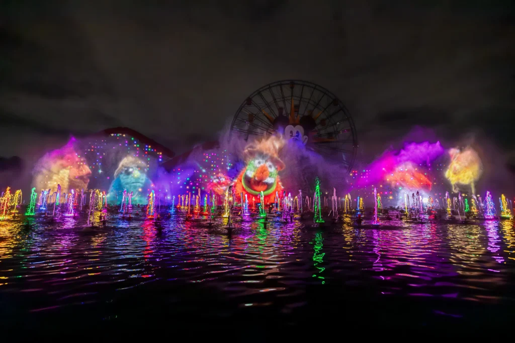 Una imagen del show World of Color Happiness, al front se van various personajes de los muppets desplegados sobre pantallas de agua, en la parte de atrás se ve la rueda de la fortuna con la cara de Mickey que se encuentra en el parque de Disney California Adventure.